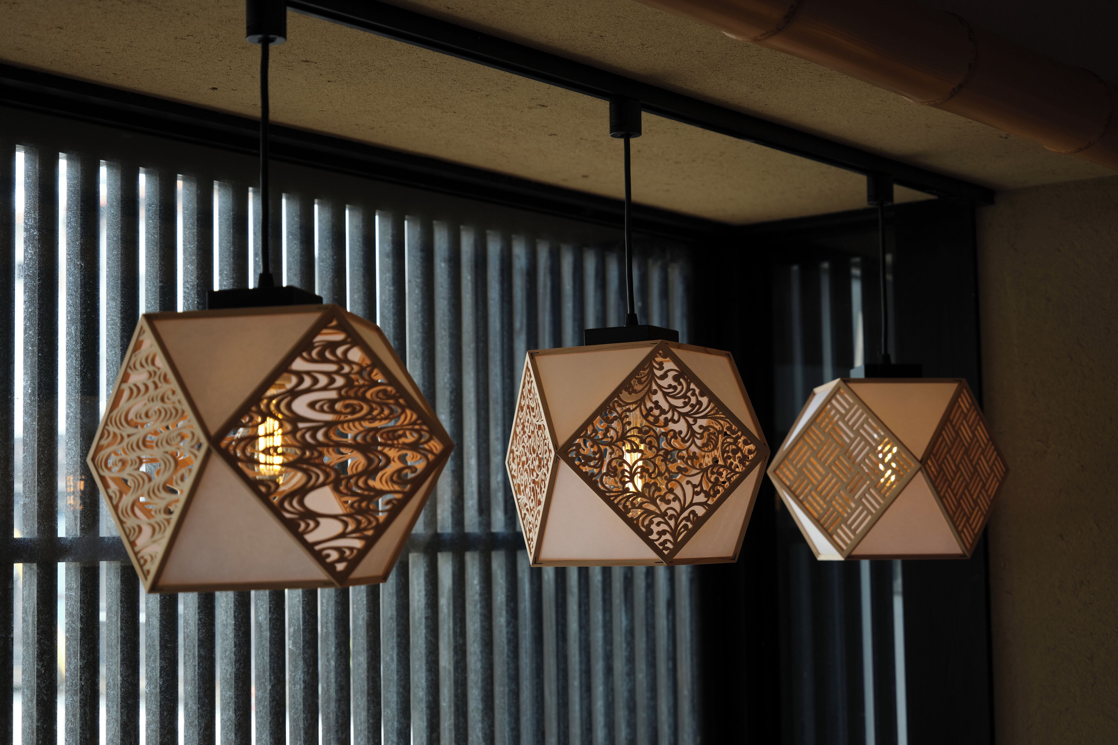 Three hexagonal hanging lights with intricate cut-out patterns, suspended from a ceiling in front of a window with vertical blinds.