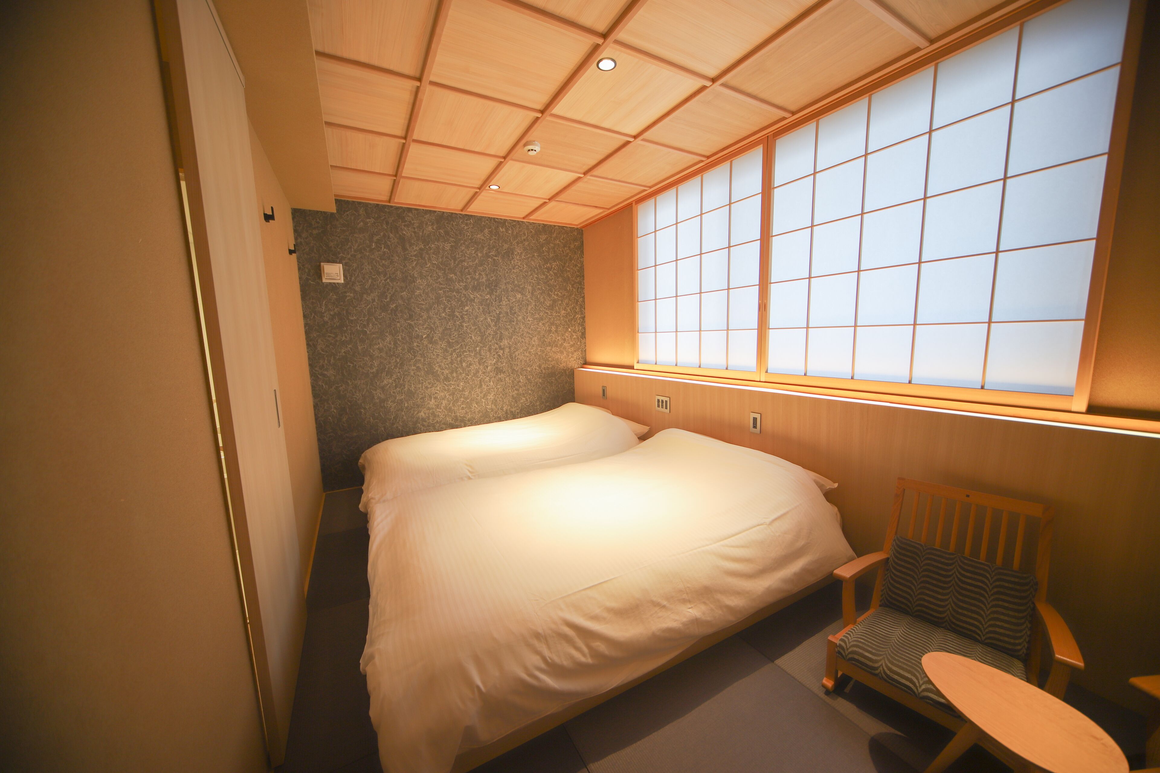 A compact hotel room with a single bed, a small wooden chair, a small table, and a large window with a view of the outside.