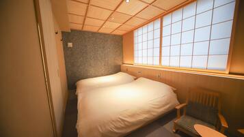 A compact hotel room with a single bed, a small wooden chair, a small table, and a large window with a view of the outside.