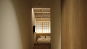 Un estrecho pasillo conduce a un dormitorio con suelo de madera, una cama con ropa blanca y una ventana con marcos de madera.