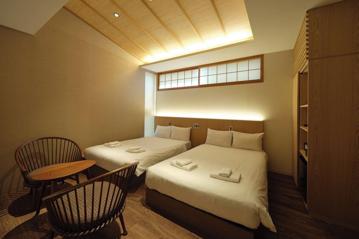 A hotel room with two beds, a wooden chair, a small table, and a wardrobe.