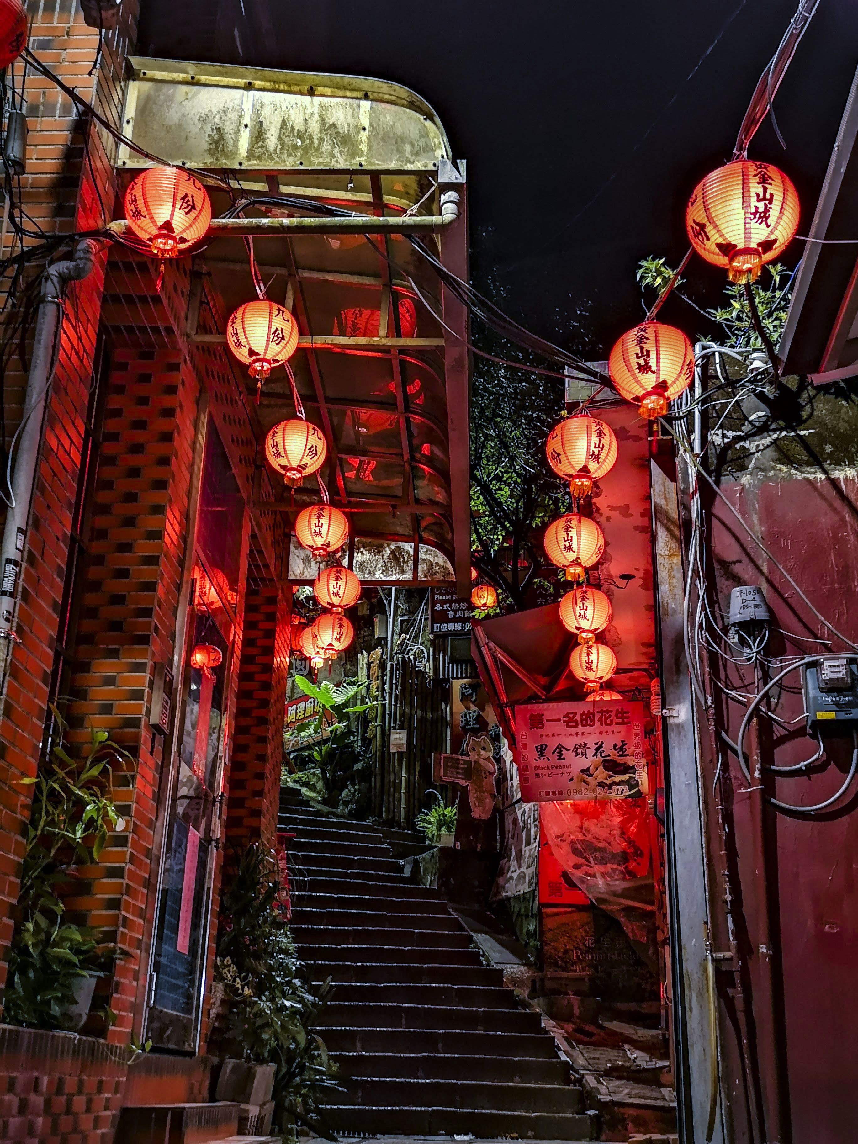 A narrow alleyway with red lanterns hanging overhead, brick walls, and a staircase leading to a higher level.