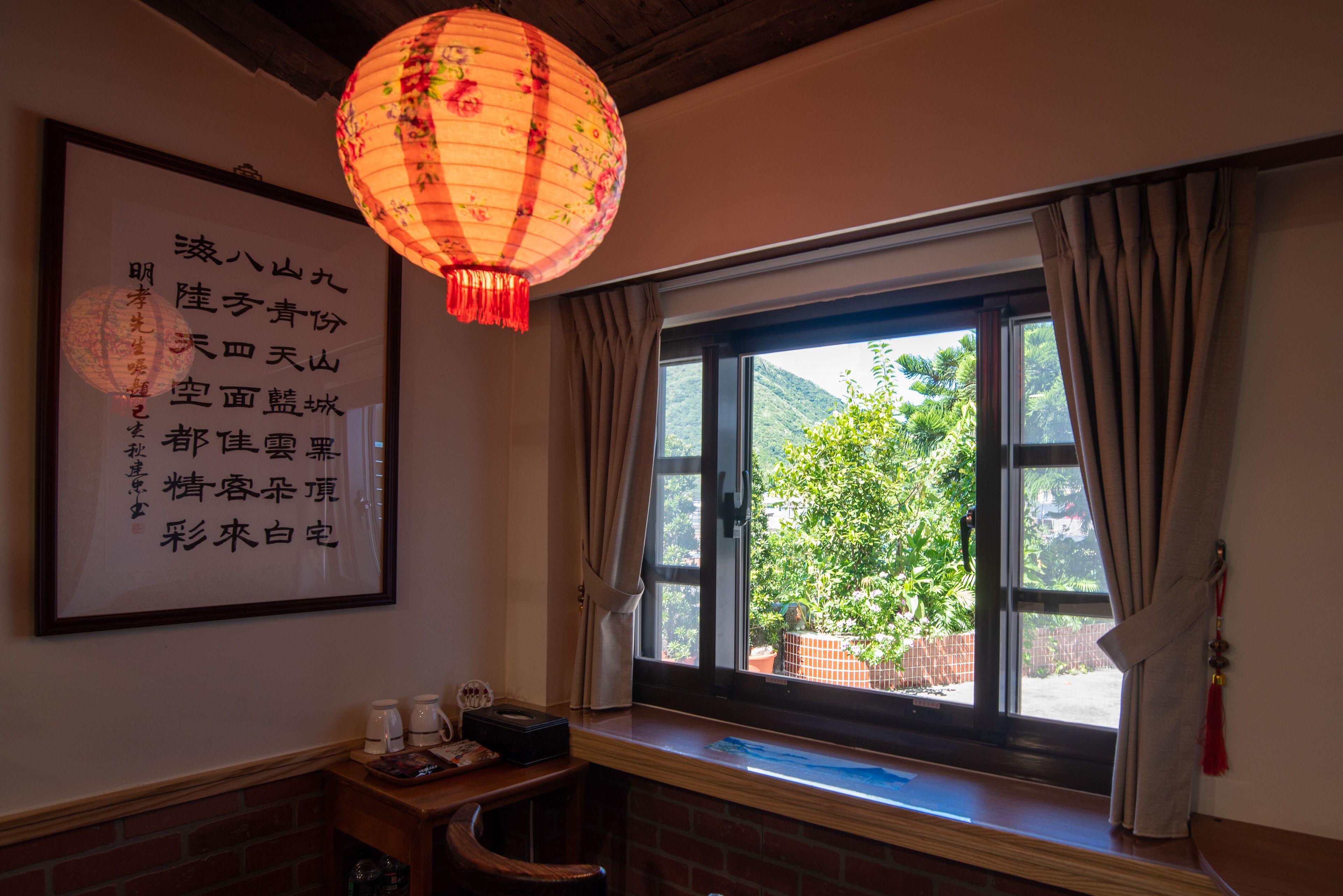 A room with a large window, a wooden bench, a framed calligraphy piece, and a hanging lantern.