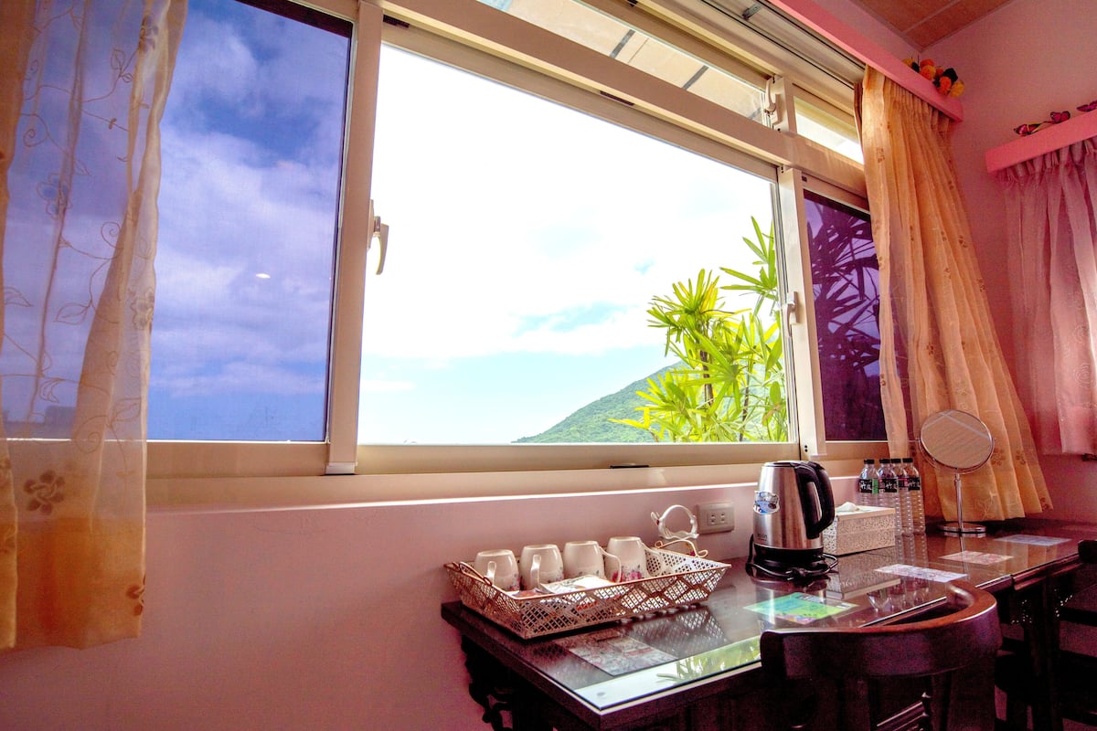 A room with a large window, a table with a kettle, cups, and bottles, and a small plant.