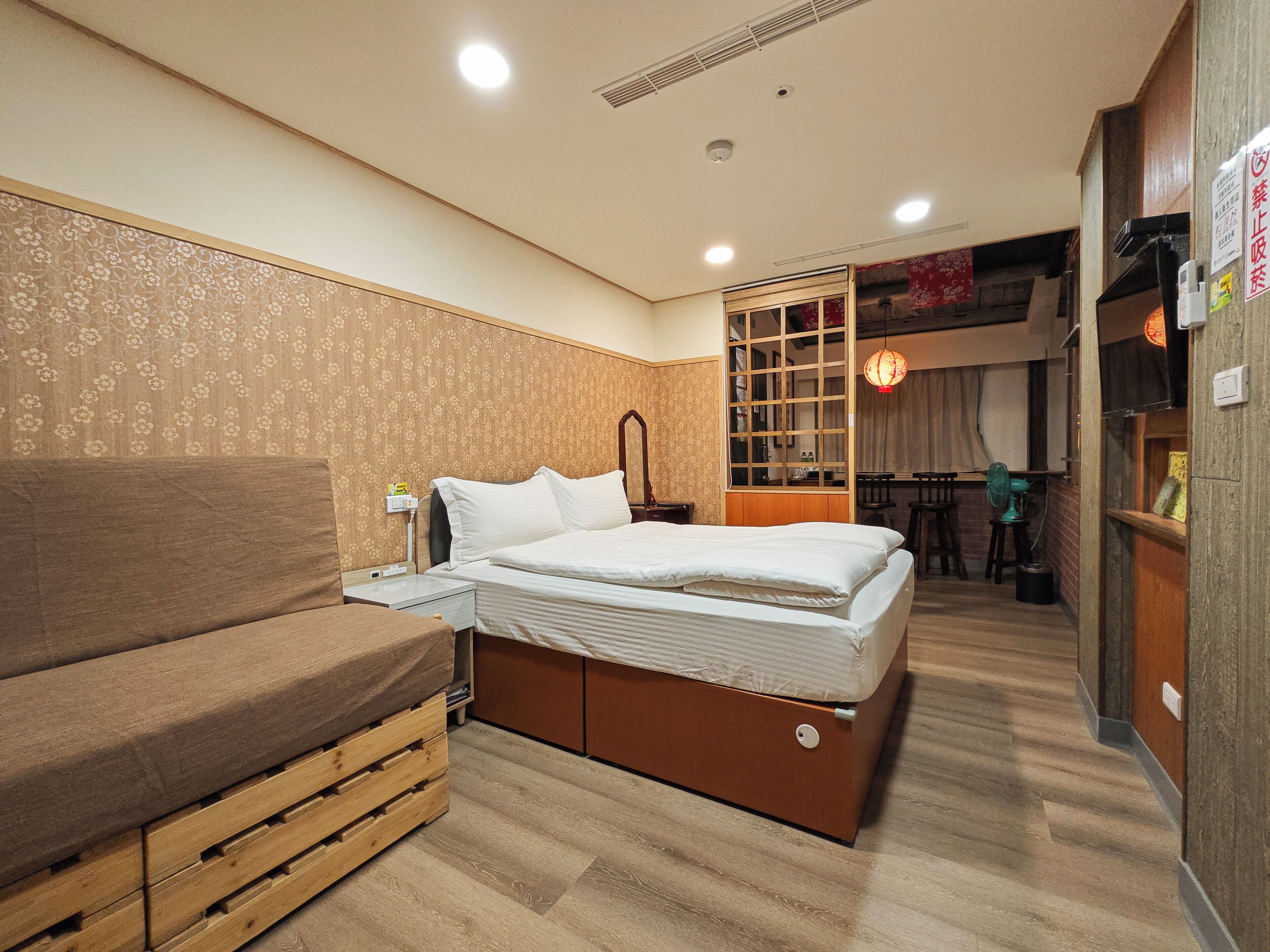 A compact hotel room with a bed, a wooden bench, a desk, and a small dining area.