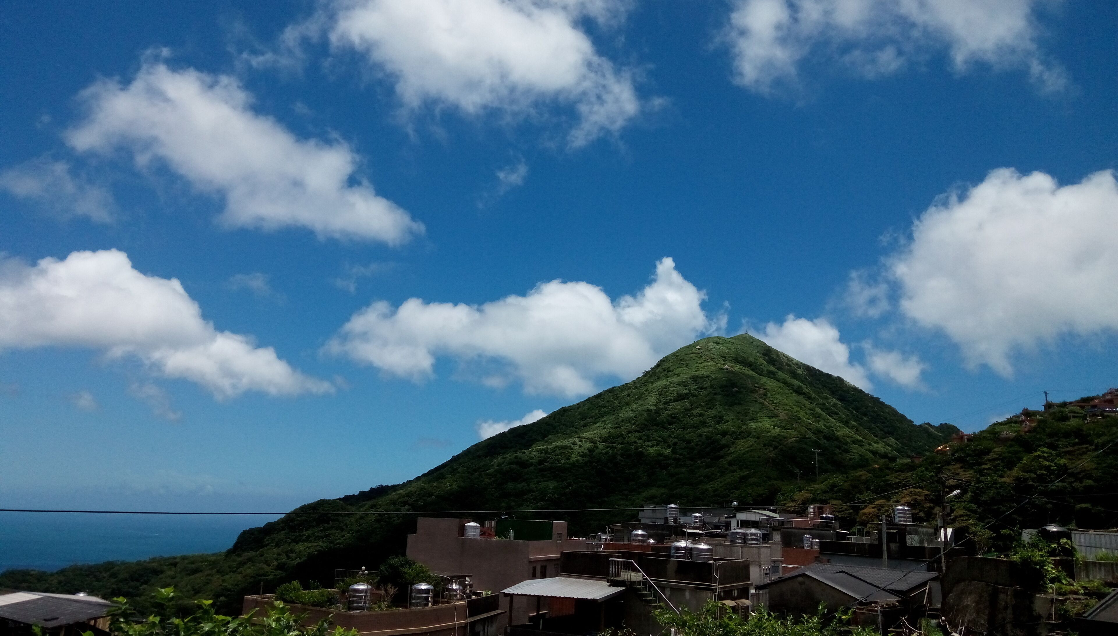 A residential area with houses on a hillside, a prominent green hill in the background, and a clear blue sky with clouds.