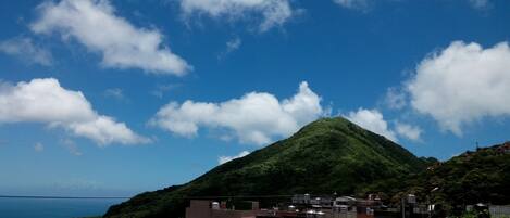 A residential area with houses on a hillside, a prominent green hill in the background, and a clear blue sky with clouds.