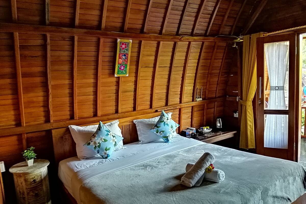 Ein Zimmer aus Holz mit einem Bett, Kissen, einem kleinen Tisch und einem Fenster mit Vorhängen.