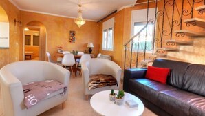 Una sala de estar con un sofá, dos sillones, una mesa de comedor con sillas, una araña y una escalera con barandillas decorativas.