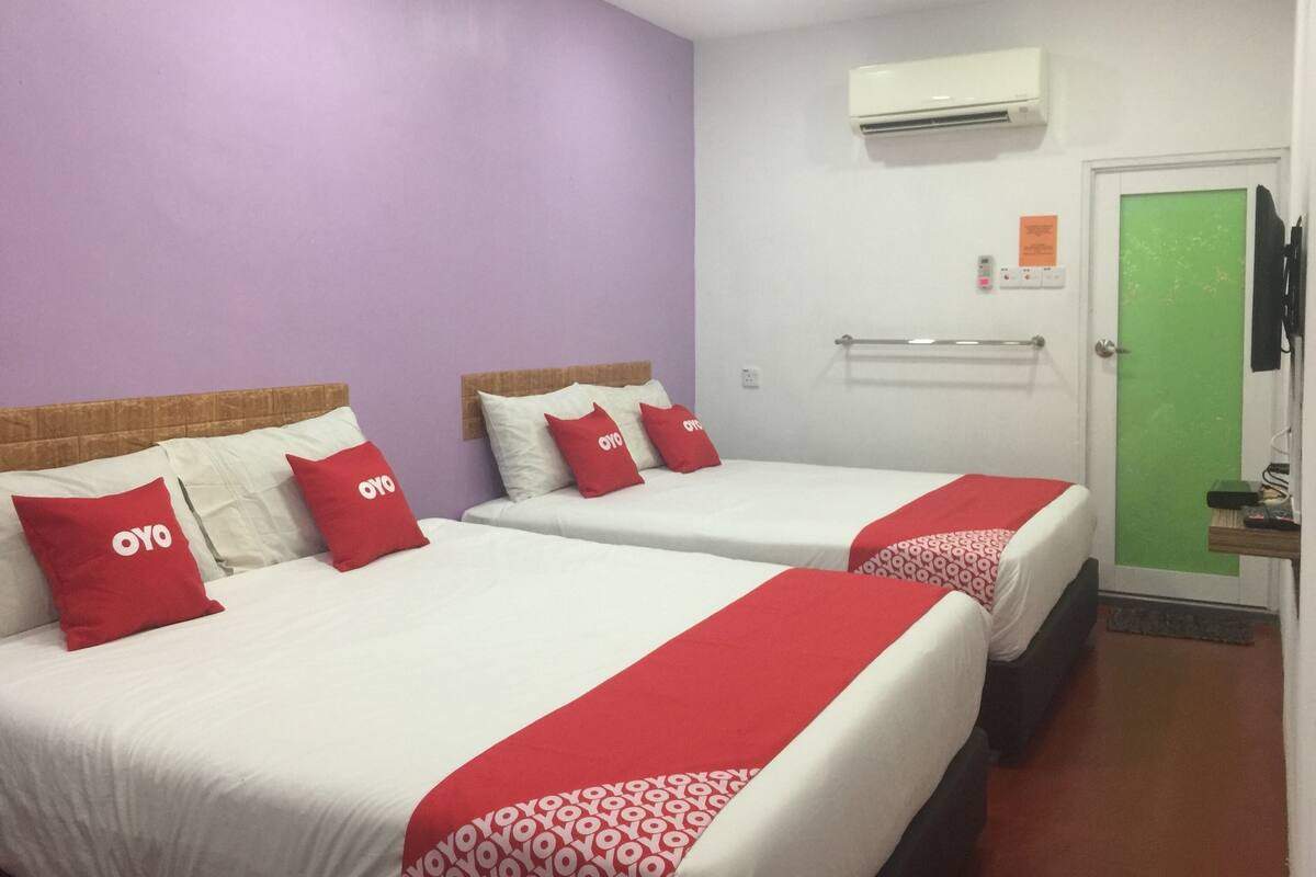 Hotelkamer met twee bedden, elk met rode en witte beddengoed en kussens, een groene deur en een airconditioner aan de muur.