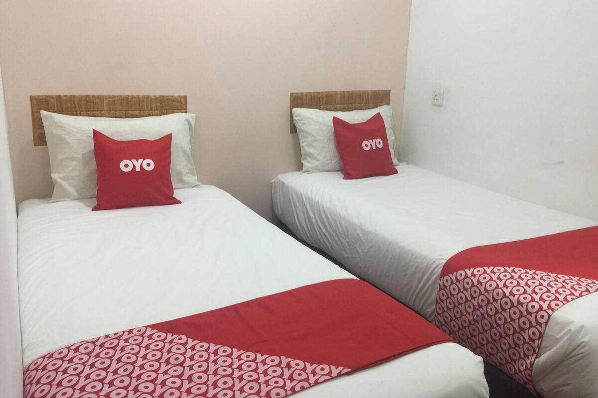 Twee enkele bedden met witte lakens en rode kussens, elk met een 'OYO'-logo, in een eenvoudige kamer met effen muren.