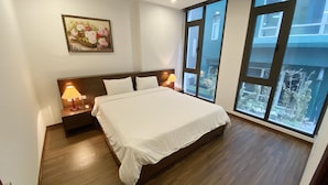A hotel room with a large bed, two bedside tables with lamps, a painting on the wall, and large windows offering a view of the outside. - Apartment Kim Ma (Hanoi)