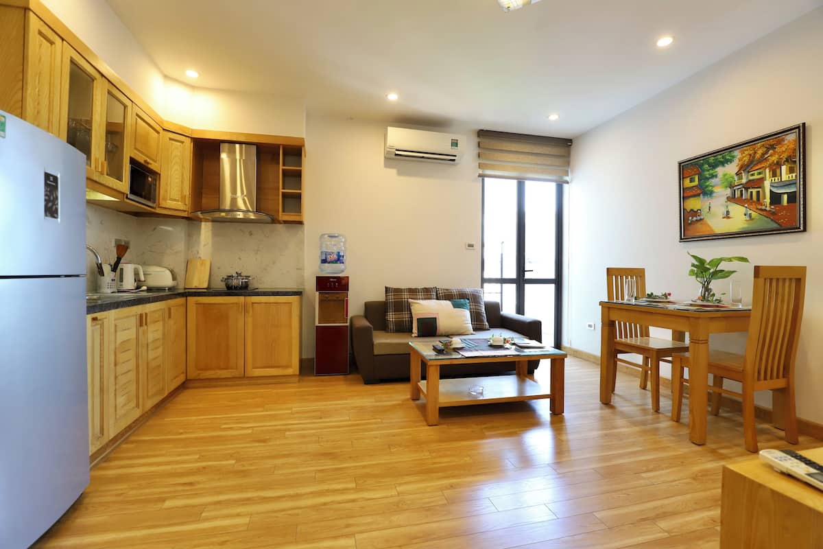 A modern kitchen with wooden cabinets, a built-in microwave, and a refrigerator. There is a dining area with a wooden table and chairs, and a sofa.