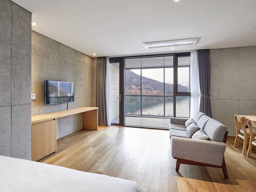 A modern hotel room with a large window, a flat-screen TV, a sofa, a desk, and a dining area.