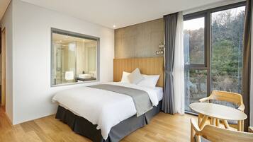 A modern hotel room with a large bed, a wooden headboard, a glass shower enclosure, and a view of trees through a large window.
