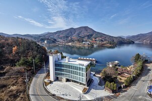 A modern building with a glass facade labeled "HAUS" near a lake, surrounded by hills and a road.