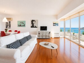 A modern living room with a white sofa, a coffee table, and a TV. There are red chairs, a dining table, and artwork on the walls. A balcony with a view of the sea is visible. - Adelos Coastal Chic by the Sea - Athens Riviera (Alimos)