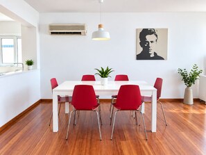 Un espace repas moderne avec une table blanche, des chaises rouges, un luminaire suspendu et un climatiseur fixé au mur.