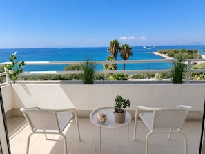 A balcony with two chairs and a small table, overlooking a coastal area with palm trees and a clear blue sky.