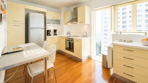 A modern kitchen with a refrigerator, oven, and coffee maker. There is a dining table with chairs and a window with a view of buildings. - Adelos Coastal Chic by the Sea - Athens Riviera (Alimos)