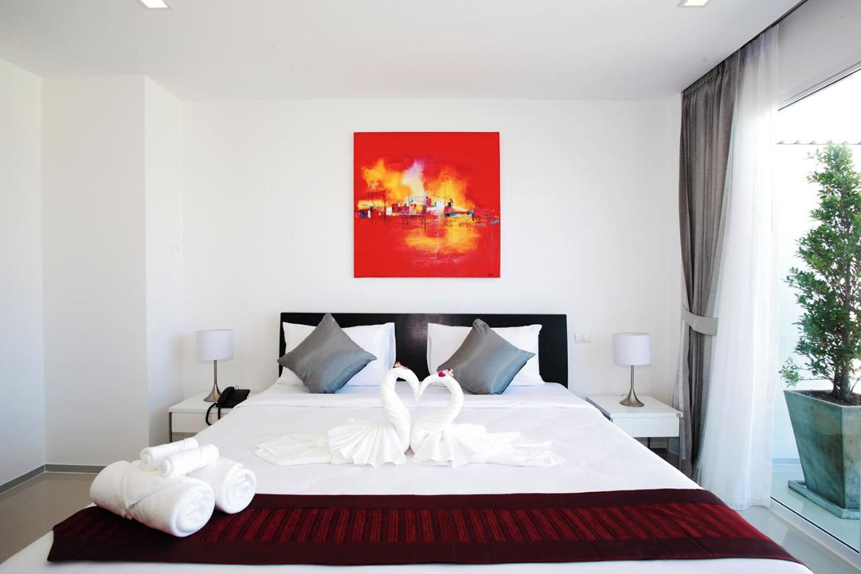 A hotel room with a large bed, two nightstands, a painting above the bed, and a potted plant by the window.