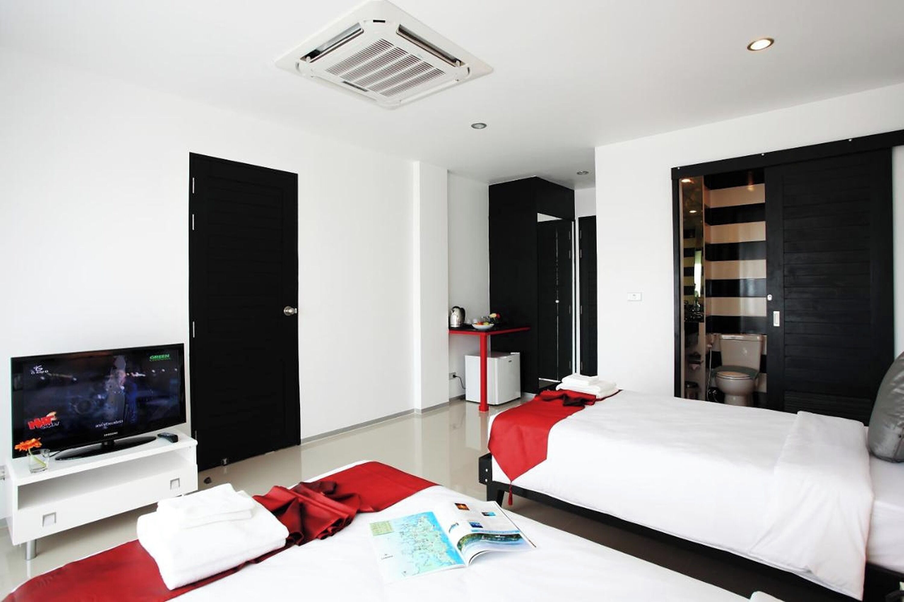 A modern hotel room with a flat-screen TV, a bed with red bedding, a white bedside table, and a red throw blanket.