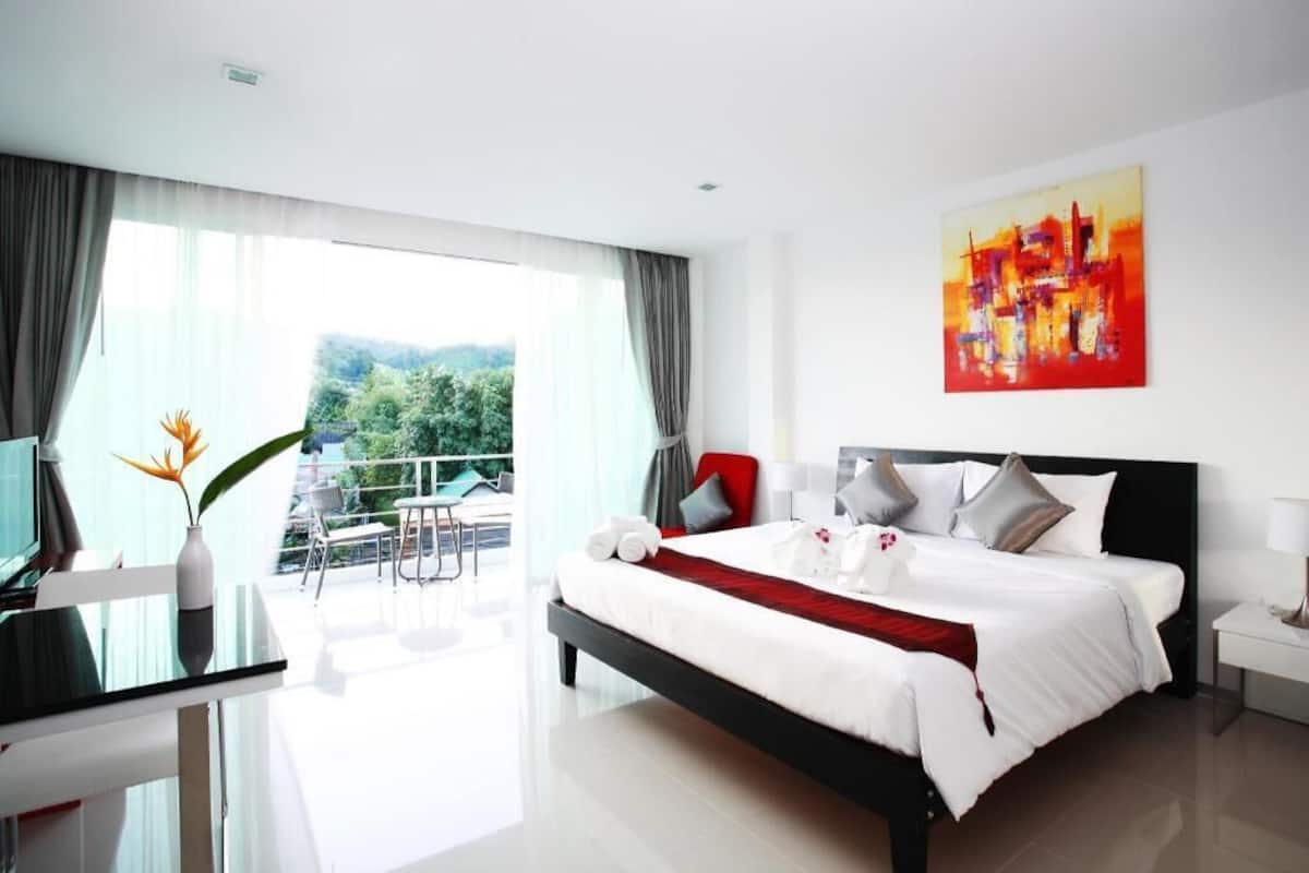 Une chambre d’hôtel moderne dotée d’un grand lit, d’un bureau avec une chaise, d’un balcon offrant une vue sur la verdure et d’un tableau au mur.