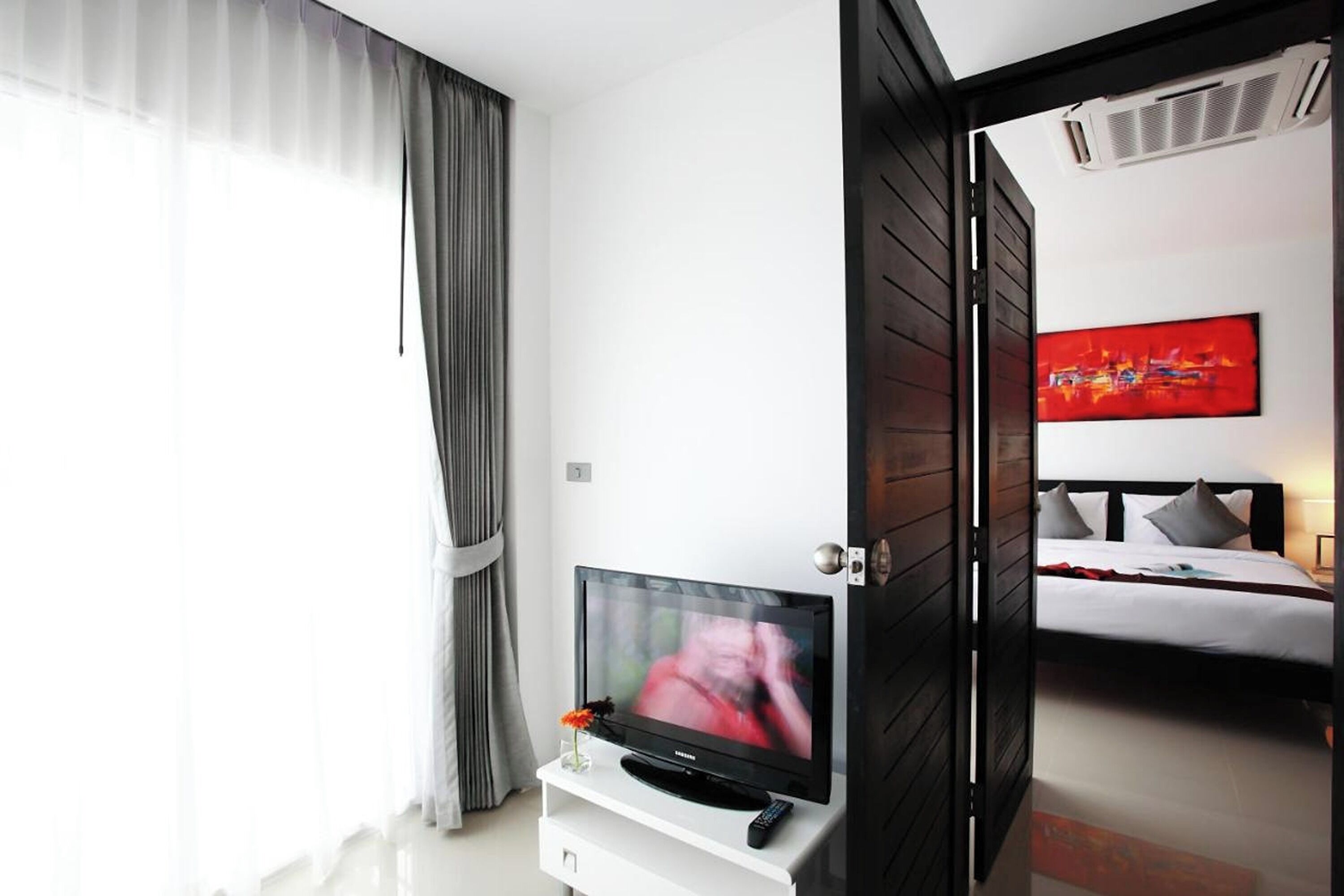 A modern hotel room with a flat-screen TV on a white stand, a bed with white bedding, a red abstract painting on the wall, and a view of the adjacent room.