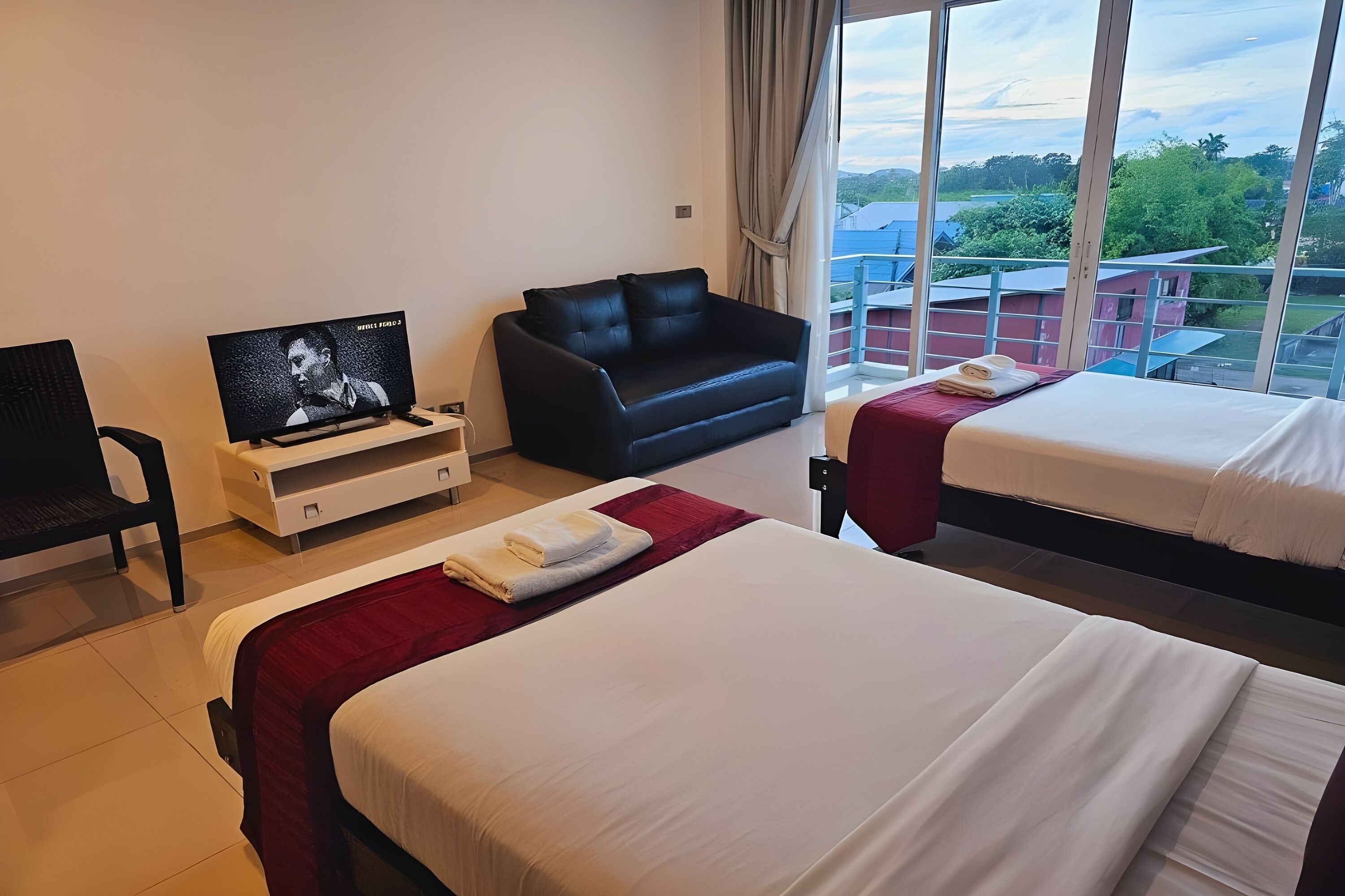 A hotel room with two beds, a TV, a sofa, and a balcony with a view of greenery.
