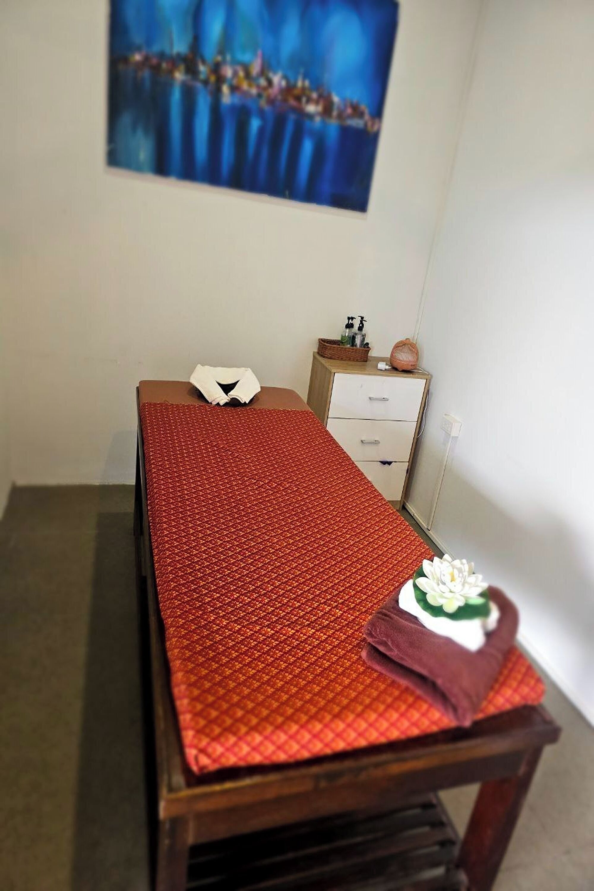 A small, neatly arranged room with a single bed, a nightstand, a small cabinet, and a wall-mounted painting.