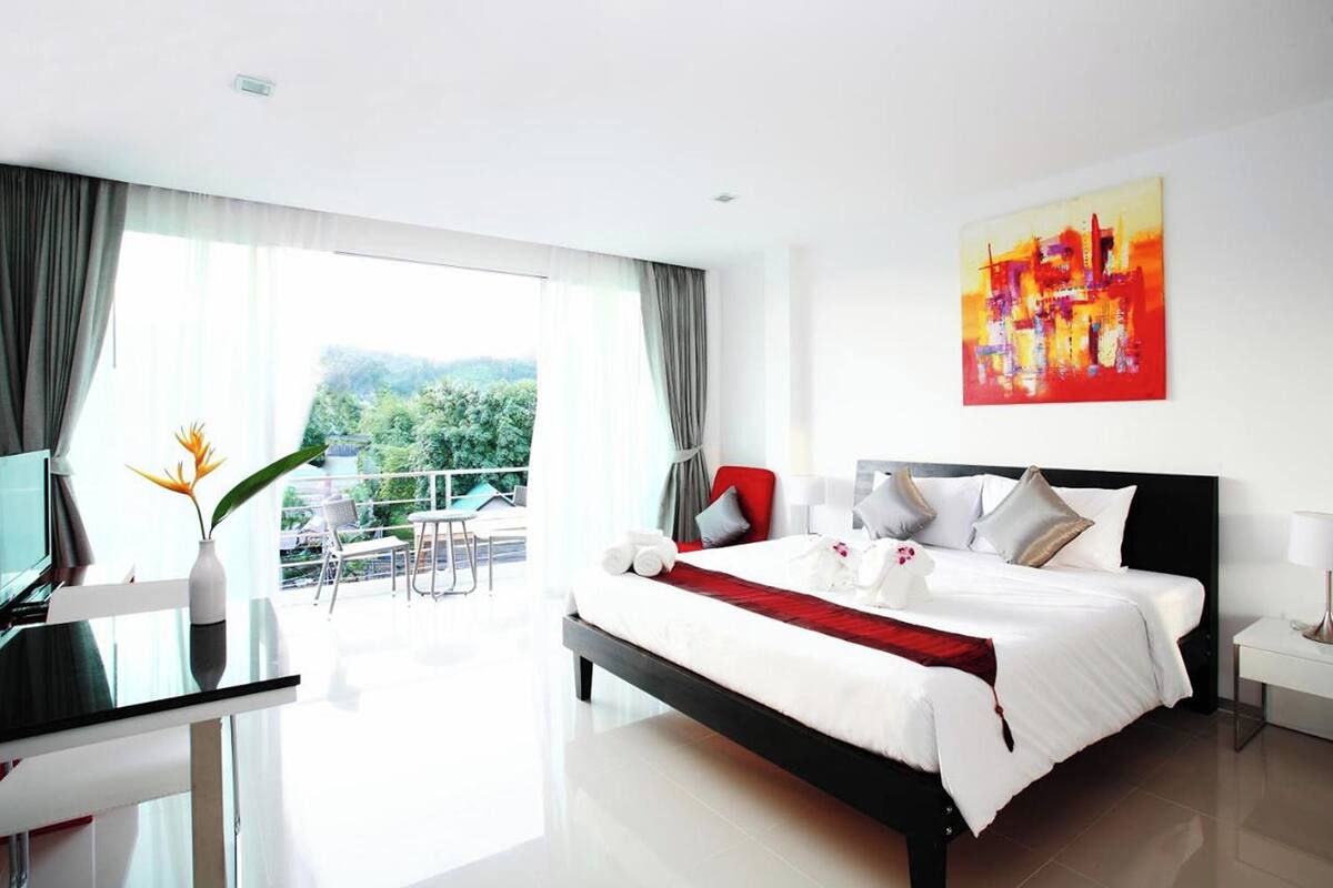 Une chambre d’hôtel moderne dotée d’un grand lit, d’un bureau avec un ordinateur et d’un balcon offrant une vue sur la verdure.