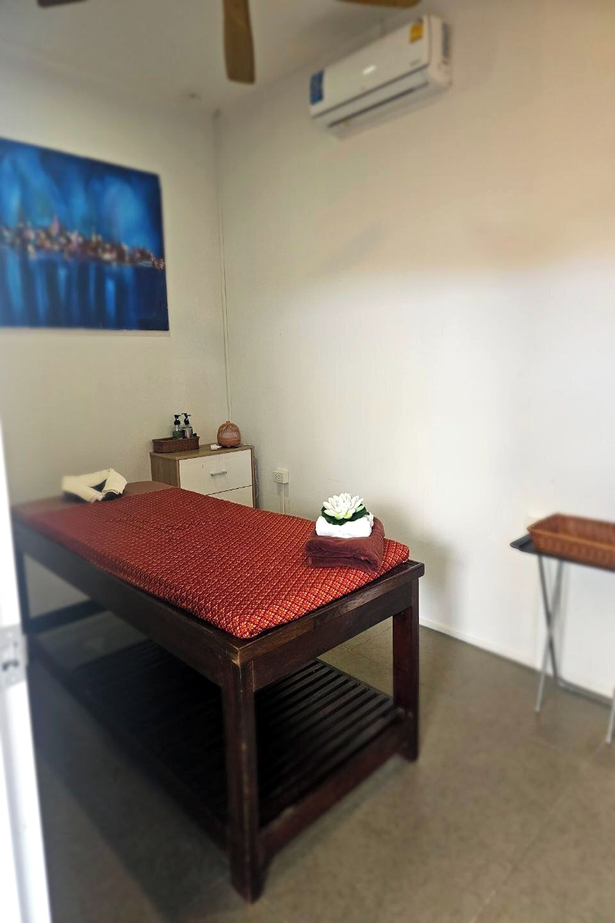 A small, clean room with a single bed, a small table, a cabinet, and an air conditioning unit.