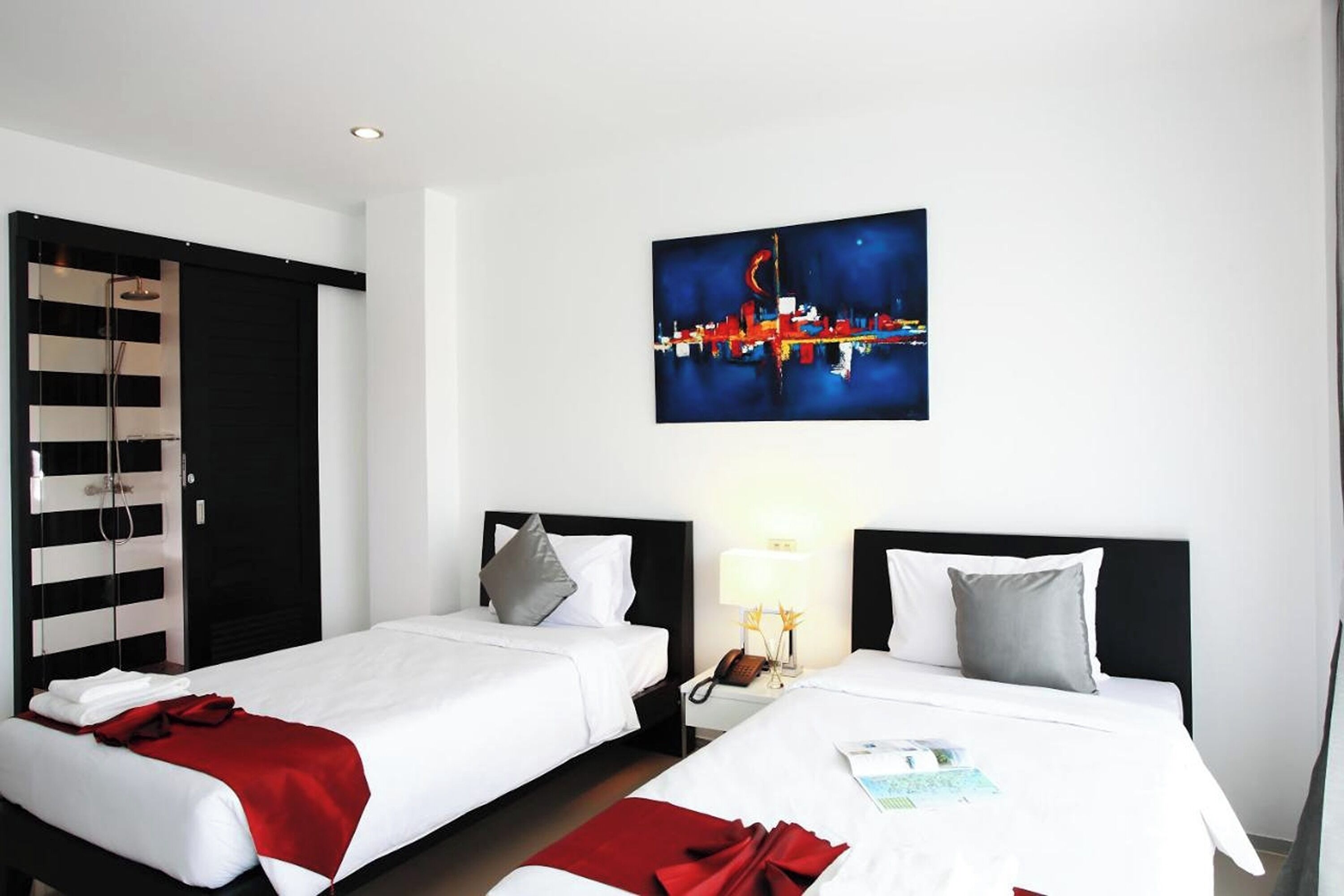 A hotel room with two beds, a cityscape painting, a nightstand with a lamp, and a black-framed window.