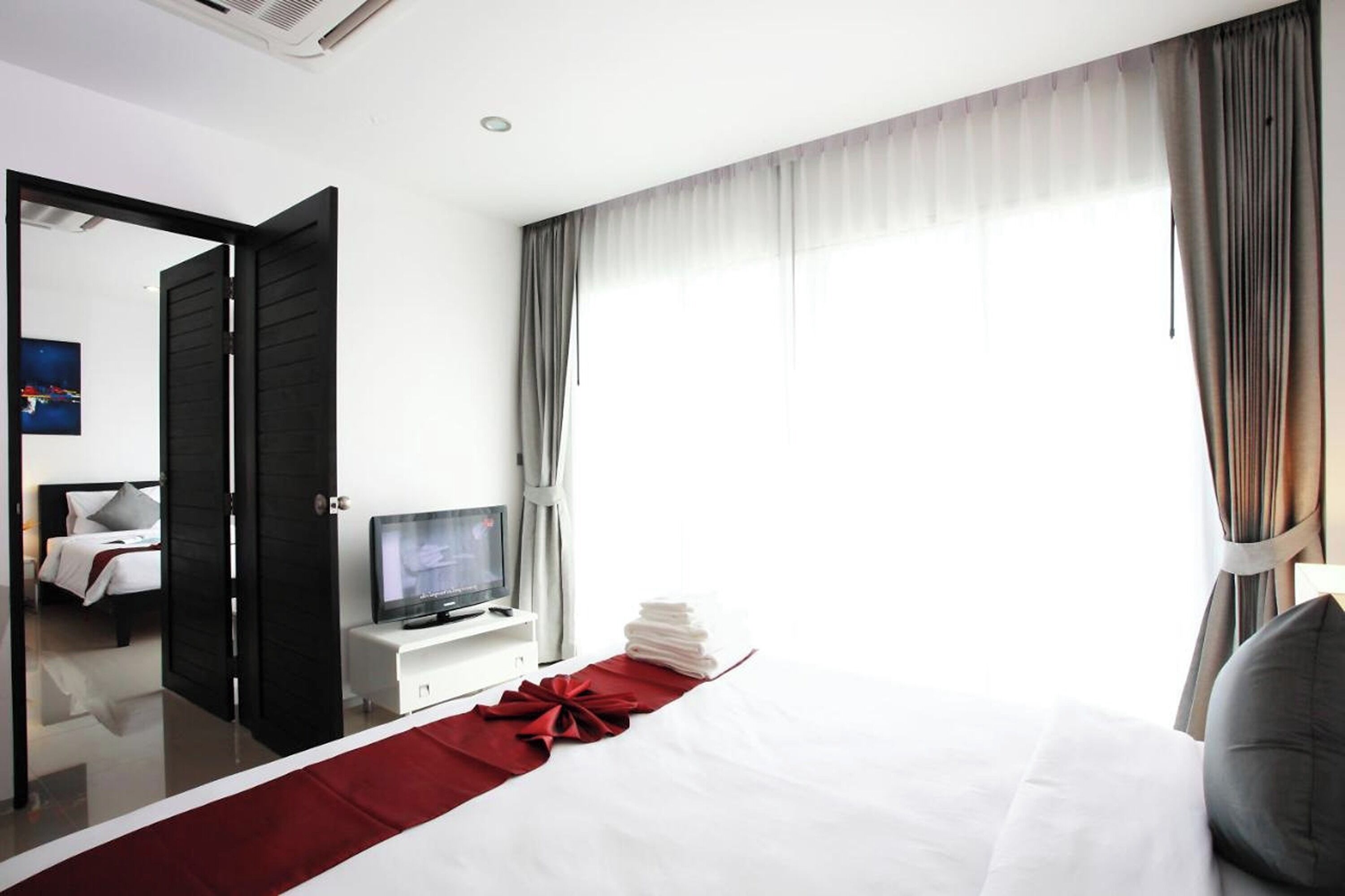 A modern hotel room with a bed, a TV on a white stand, a red runner, and a view of the outside through large windows.