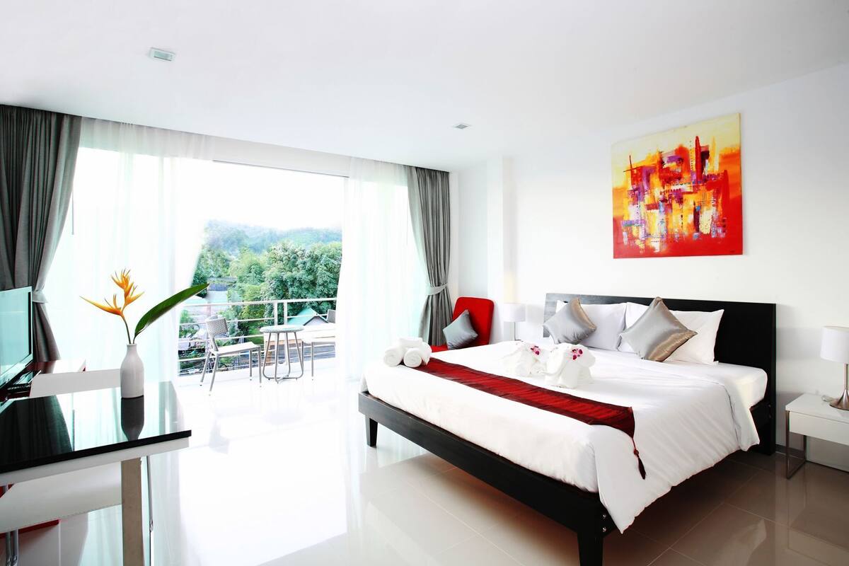 Une chambre d’hôtel moderne, dotée d’un grand lit, d’une télévision, d’un balcon offrant une vue sur la verdure et d’un tableau au mur.