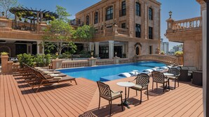 A rooftop pool area with lounge chairs, tables, and a large building with balconies.