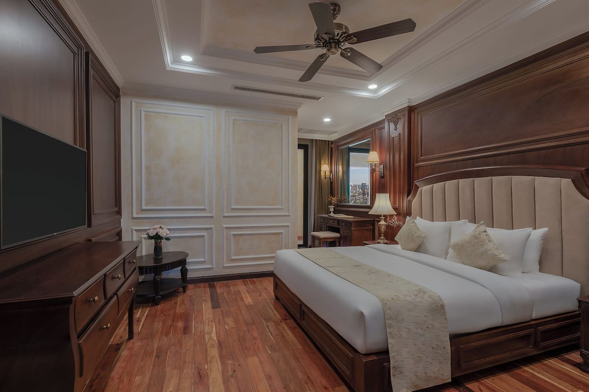Quarto de hotel com uma cama grande, móveis de madeira, ventoinha no teto e vista para a cidade pela janela.