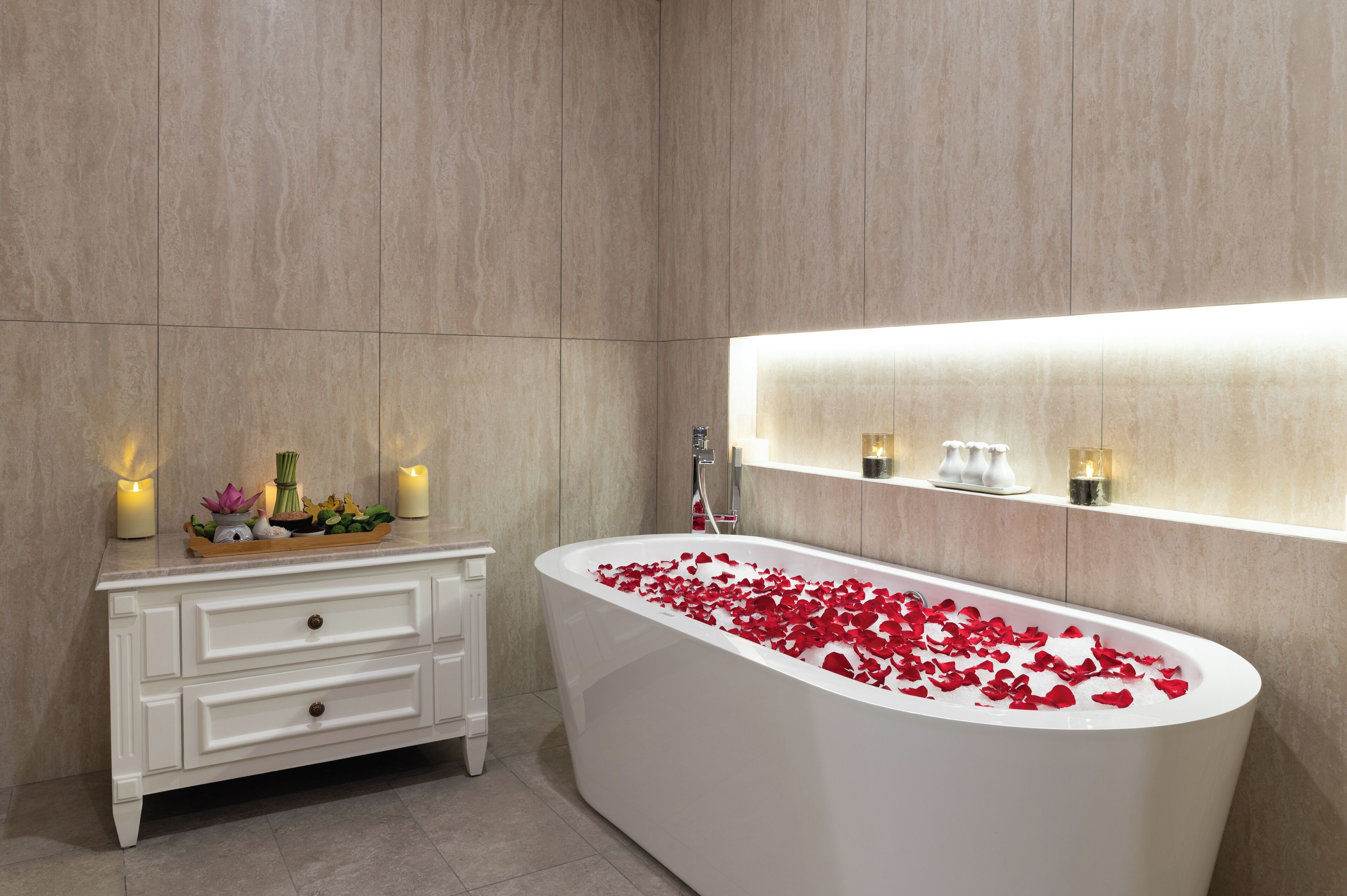 A modern bathroom with a freestanding bathtub filled with rose petals, a white vanity with drawers, and wall-mounted sconces.