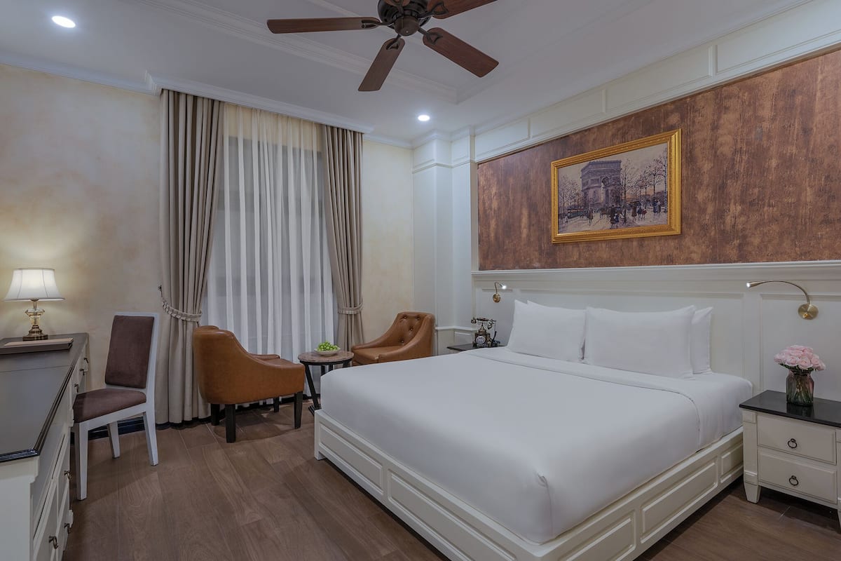Quarto de hotel com uma cama grande, uma escrivaninha com luminária, duas cadeiras, um ventilador de teto e um quadro na parede.