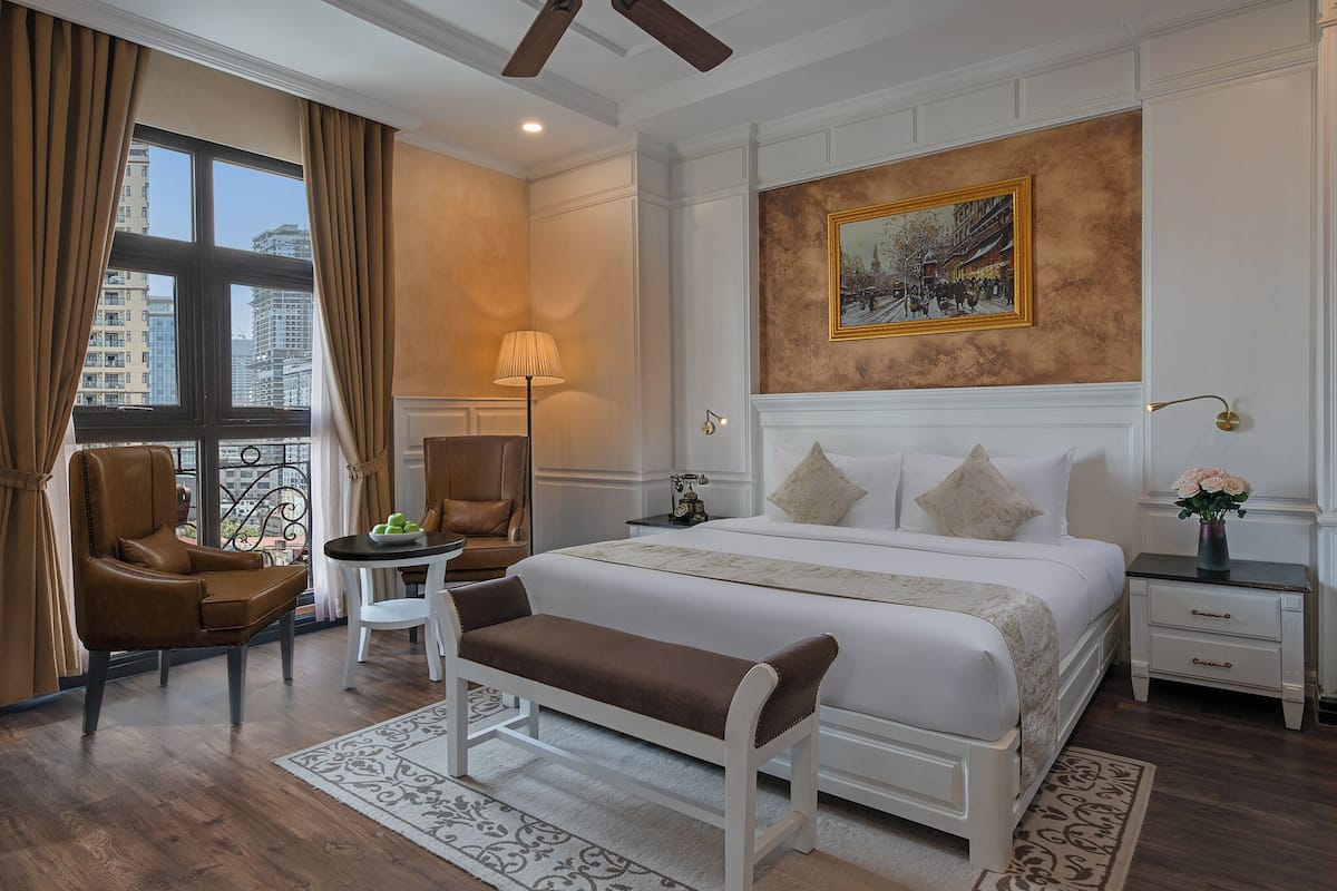 Quarto de hotel com uma cama grande, um banco, uma cadeira, uma mesa pequena, uma luminária, uma mesinha de cabeceira e um quadro na parede.