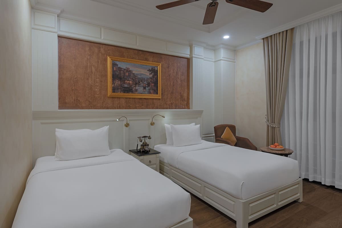 Quarto de hotel com duas camas, um quadro na parede, um ventilador de teto e uma janela com cortinas.