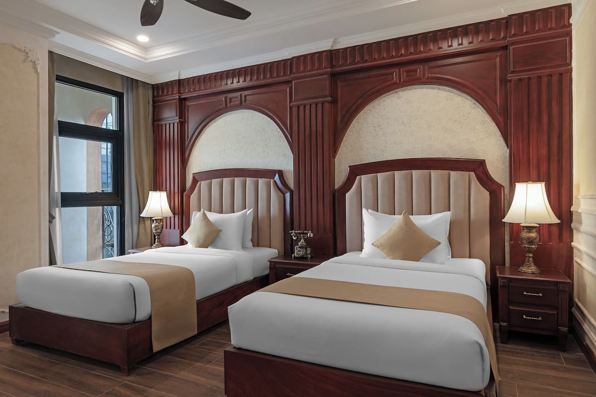 Quarto de hotel com duas camas, cabeceiras de madeira e uma grande janela em arco.