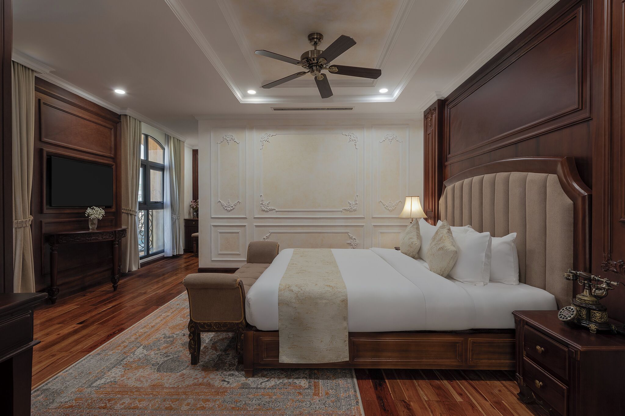 A large, elegantly furnished bedroom with a wooden bed, a ceiling fan, and a television on a wooden stand.