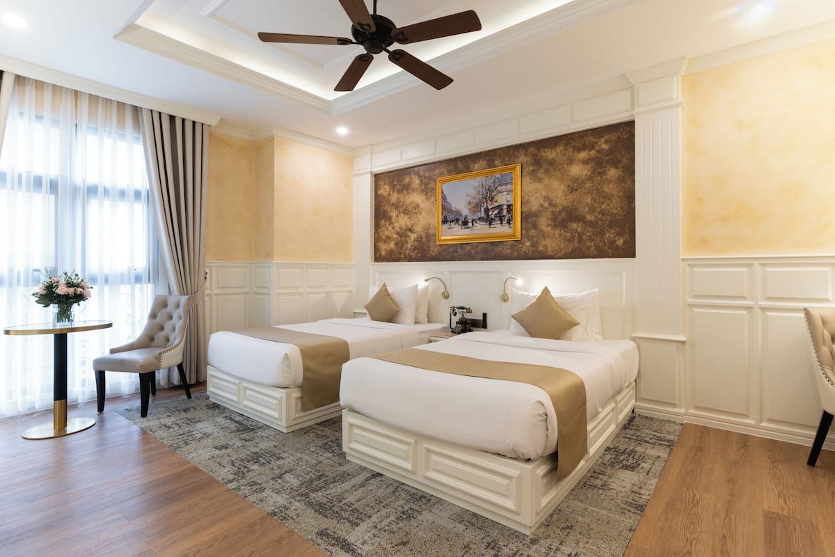 Quarto de hotel com duas camas, um ventilador de teto, um quadro na parede e uma pequena mesa com um vaso de flores.