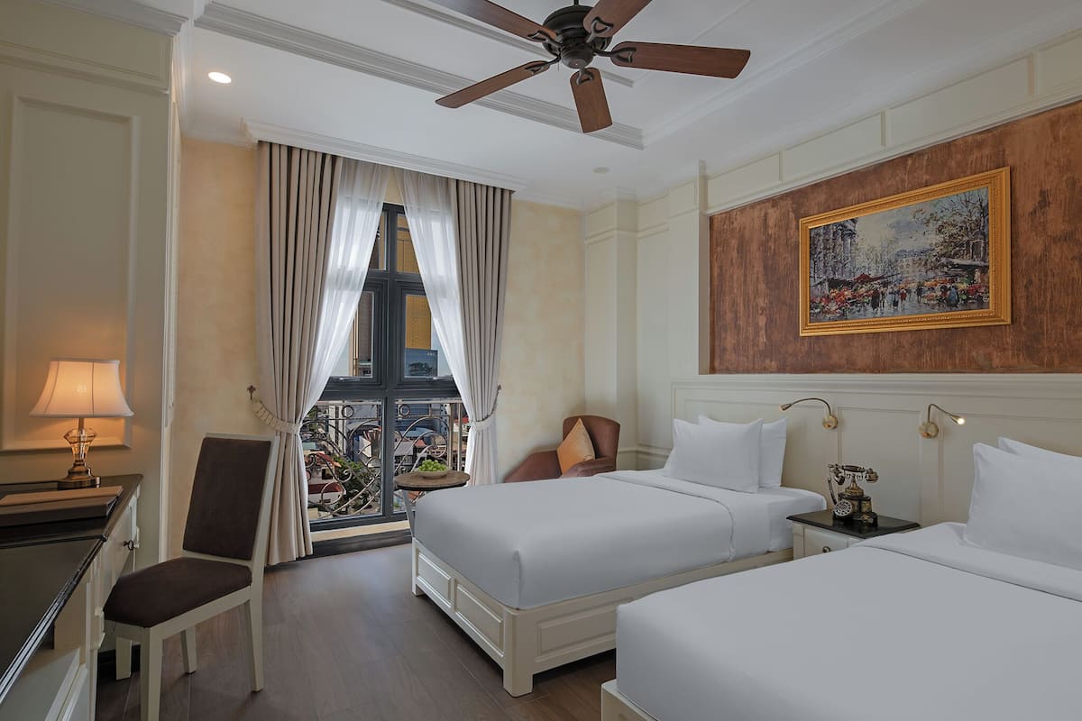 Quarto de hotel com duas camas, uma escrivaninha, uma cadeira, um ventilador de teto, uma janela com cortinas e um quadro na parede.