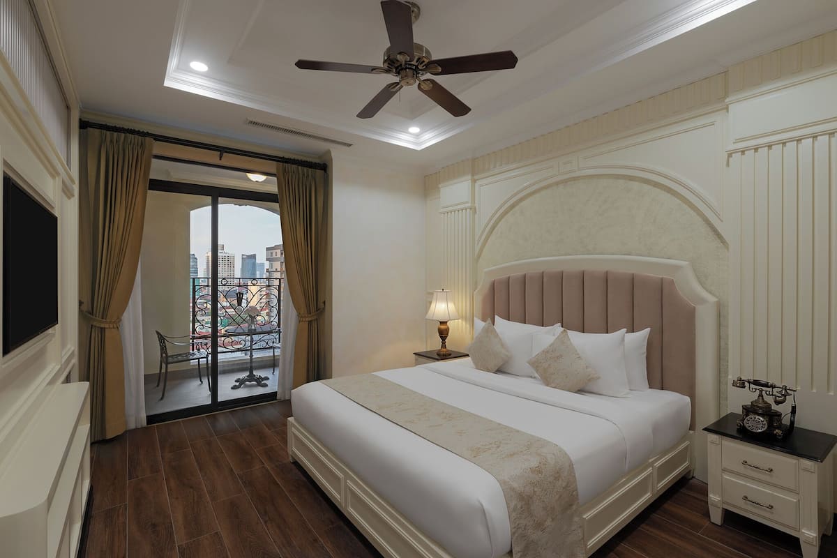 Quarto de hotel com uma cama grande, um ventilador de teto, uma mesa de cabeceira com abajur e vista da cidade através de uma porta de varanda.