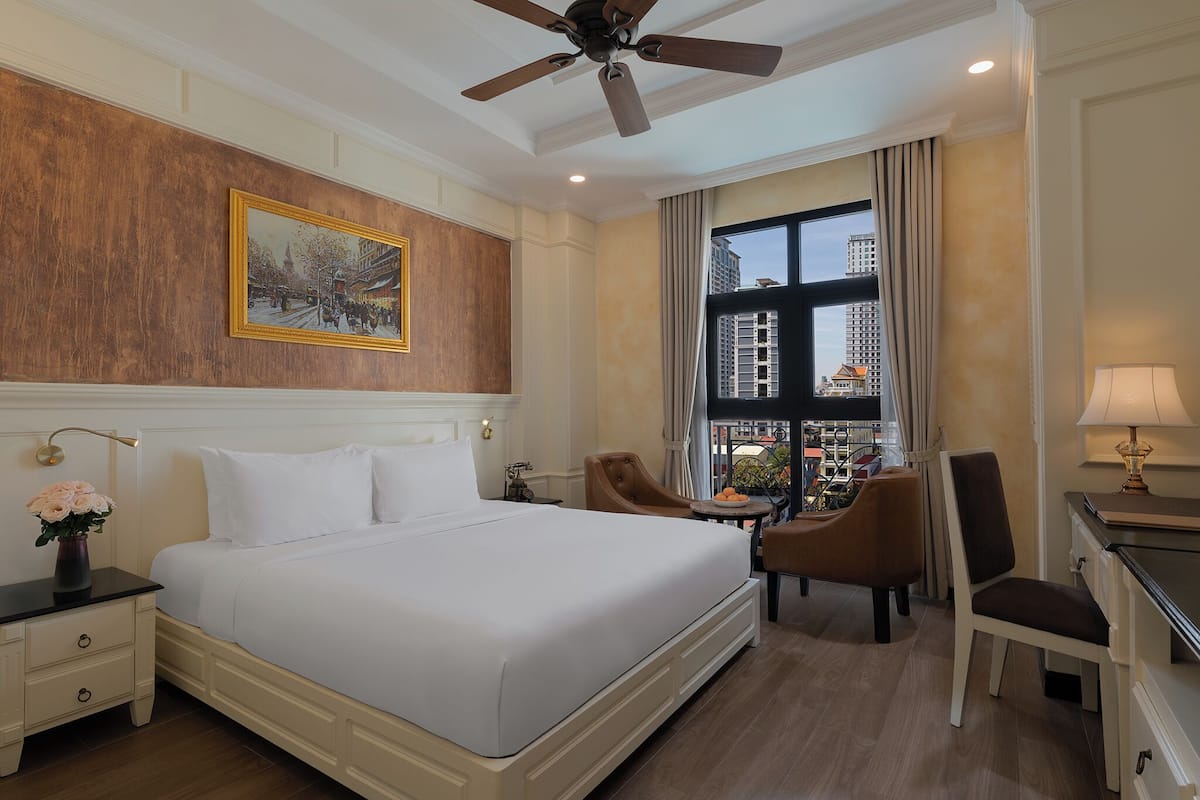 Quarto de hotel com uma cama grande, uma escrivaninha, duas cadeiras, um ventilador de teto, uma janela com vista para a cidade e um quadro na parede.