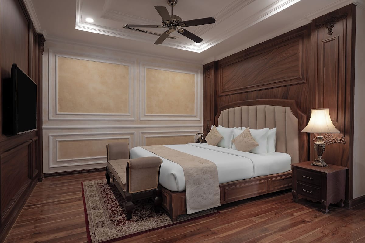 Quarto com uma cama grande, ventoinha no teto, televisão, mesa de cabeceira com abajur e piso de madeira.