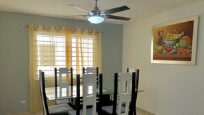 A dining area with a glass table and chairs, a ceiling fan, and a painting of fruits on the wall. - 3 Bedroom Apartment with Resort like Pool (Stgo)