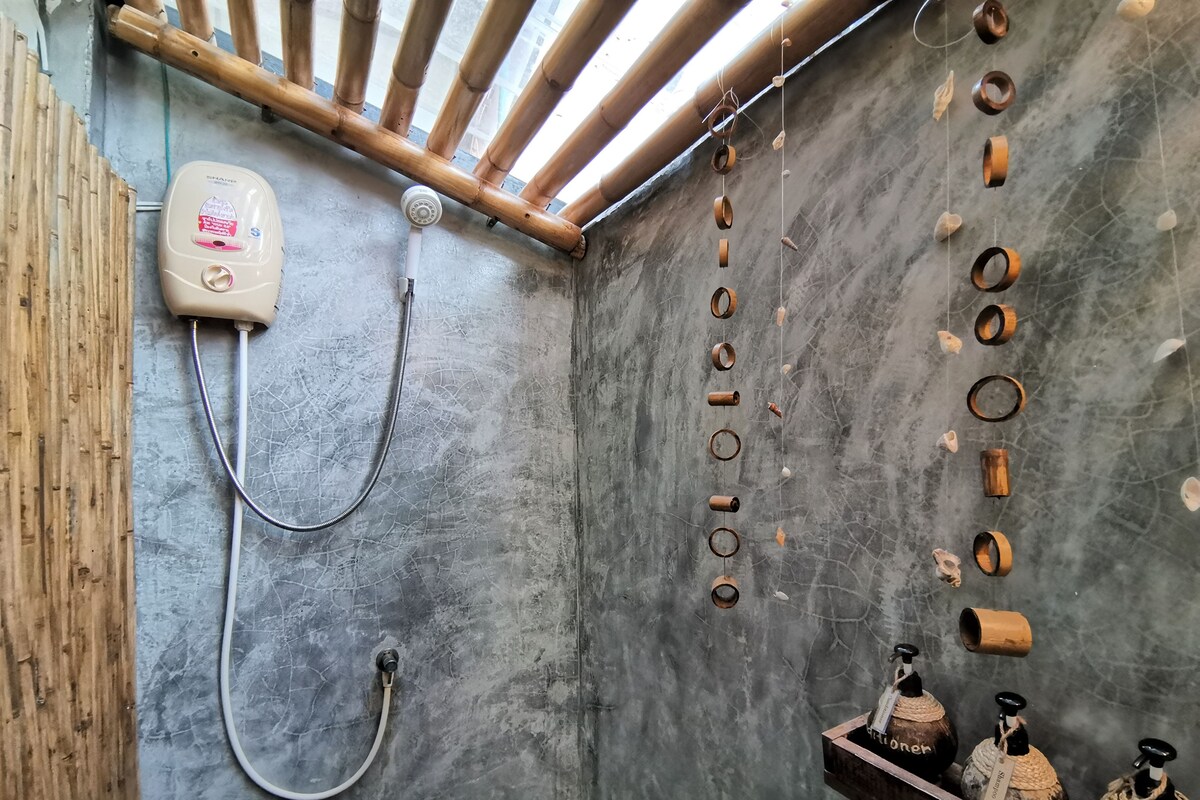 Casa de banho com parede de betão, chuveiro com barra de duche e prateleira de madeira com garrafas.